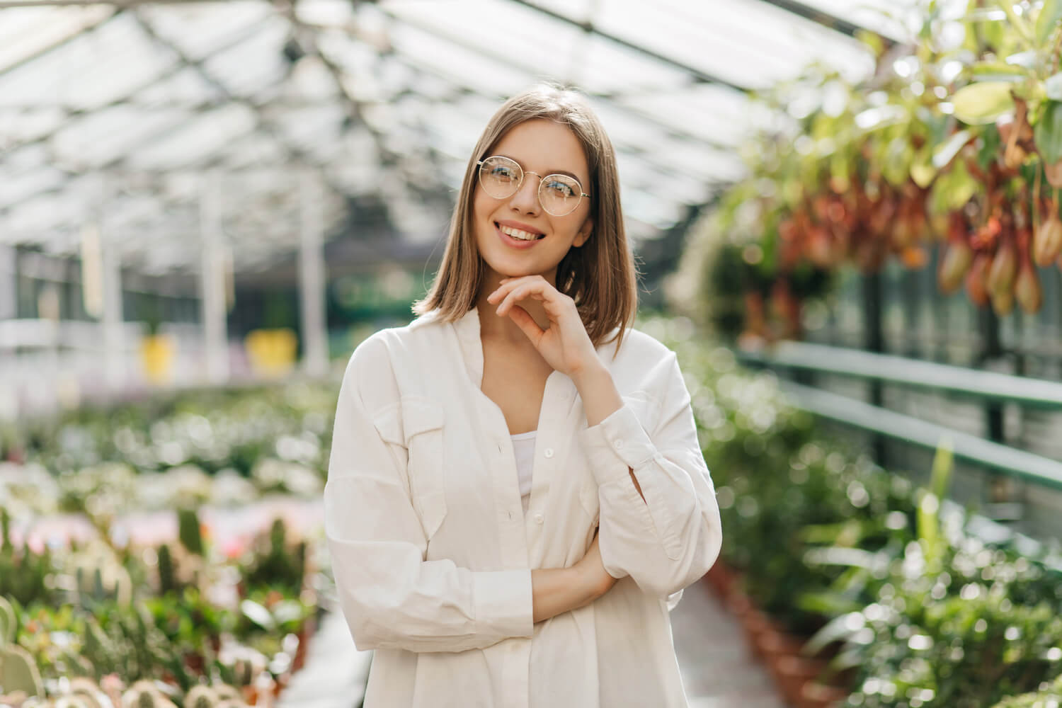 A smiling woman with glasses and shoulder-length hair stands in a greenhouse, surrounded by hanging and potted plants. She is wearing a white blouse and has her hand under her chin. The greenhouse is bright and filled with greenery.